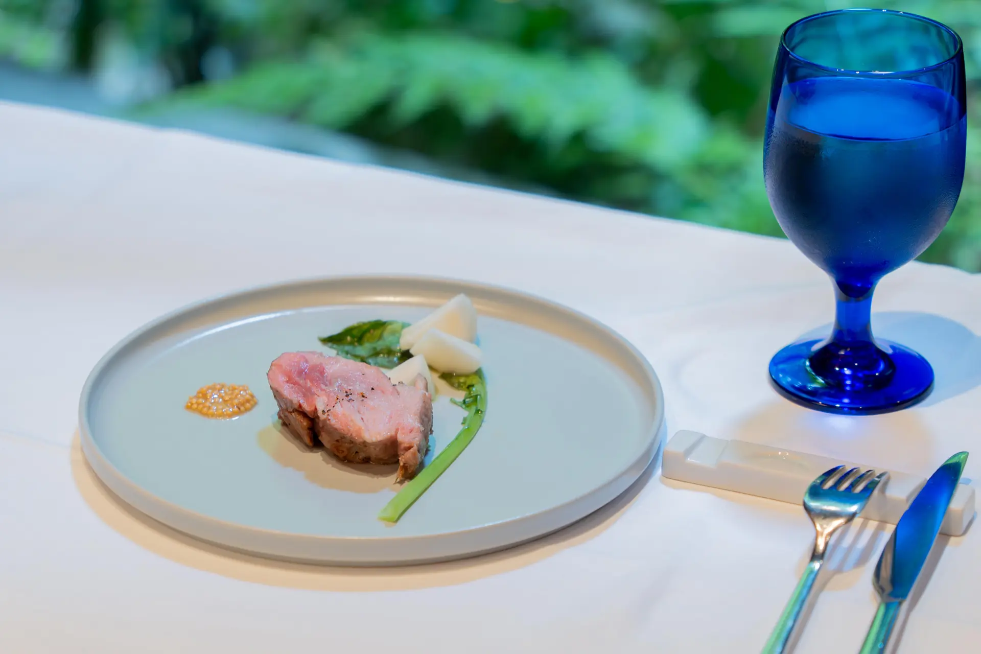 緑を背景に白いテーブルへ盛り付けられた料理と青いグラス